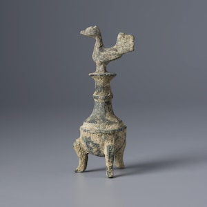 Pode incluir: Um queimador de incenso de bronze desgastado com uma figura semelhante a um pássaro no topo de um pedestal alto e ornamentado. A base de três pernas suporta um corpo bulboso. A peça tem uma pátina marrom esverdeada, sugerindo idade. O fundo é cinza liso.