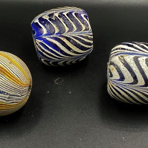 Puede incluir: Tres cuentas de vidrio con un patrón rayado. Las cuentas son azul y blanco, azul y blanco y amarillo y blanco.