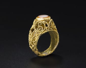 May include: A gold ring with an oval, light-colored stone. The ring features intricate filigree and beaded detailing around the band and setting. The ring is set against a black background, highlighting its ornate design.