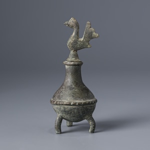 Pode incluir: Um queimador de incenso em estilo antigo, cor bronze, com um remate em forma de pássaro. O queimador tem um corpo bulboso, uma faixa decorativa e três pernas curtas. O pássaro tem um pescoço longo e uma cauda detalhada.