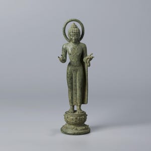 Pode incluir: Estátua de bronze de um Buda em pé com auréola. A figura está vestida com vestes e está sobre um pedestal detalhado. A estátua tem uma expressão serena e mãos estendidas, com uma pátina verde clara. Cerca de 30 cm de altura.