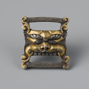 May include: A detailed, square-shaped metal object with a handle, featuring a stylized face with gold accents. The face has a prominent nose, open mouth, and detailed eyes. The metal has a dark, aged appearance.