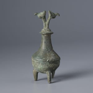 Pode incluir: Um vaso de bronze com um corpo bulboso e um pescoço fino, suportado por três pernas curtas. O topo apresenta um elemento decorativo com duas figuras semelhantes a pássaros. O vaso tem uma aparência envelhecida.