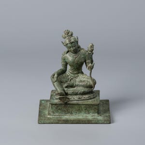 Puede incluir: Estatua de bronce de una figura sentada con detalles ornamentados, posiblemente una deidad. La figura está adornada con joyas y sostiene una flor de loto. La estatua está montada sobre una base cuadrada y tiene un aspecto envejecido.