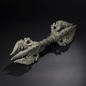 May include: An antique bronze vajra, a ritual object with three prongs on each end. The central shaft is cylindrical with a ridged texture. The ends feature intricate, stylized dragon or mythical creature heads.
