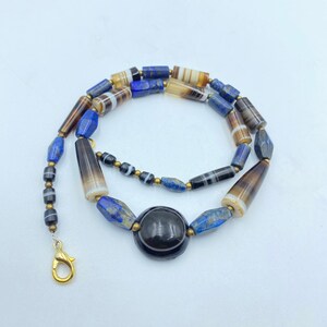 Op de afbeelding: Een ketting gemaakt van verschillende gekleurde kralen, waaronder blauwe lapis lazuli, bruine agaat en zwarte onyx. De ketting heeft een grote, ronde zwarte onyx hanger.