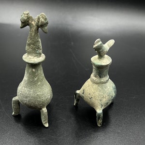 Pode incluir: Dois tampões de garrafa antigos de bronze, cada um com três pernas. Um tampão apresenta um desenho de pássaro, enquanto o outro apresenta um desenho de pássaro de duas cabeças.