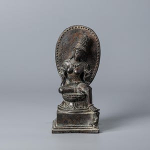 Può includere: Una statua in bronzo di una divinità seduta con un'elaborata acconciatura e gioielli. La figura è posizionata di fronte a uno sfondo circolare e strutturato. La statua è montata su una base quadrata.