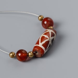 Peut inclure: Un fil de perles. Il présente une perle cylindrique brun-rouge avec un motif géométrique blanc, flanquée de deux perles rondes brun-rouge et d'entretoises dorées. Les perles sont enfilées sur un fin cordon blanc.