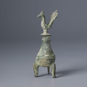 May include: A weathered bronze sculpture of a bird atop a three-legged vessel. The bird has outstretched wings and a detailed head. The vessel has a bulbous body and a flared base, showcasing an aged, green patina.