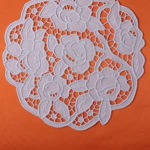 Könnte beinhalten: Ein weißes, kreisförmiges Deckchen mit floralem Design, das Rosen und Blätter zeigt. Das Deckchen besteht aus einem zarten, offen gewebten Stoff und ist vor einem orangefarbenen Hintergrund platziert. Dieser Dekorationsartikel eignet sich für die Inneneinrichtung.
