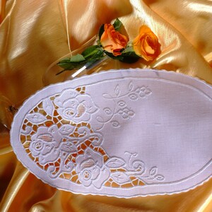 Puede incluir: Doily de encaje blanco ovalado con un delicado diseño floral con rosas y enredaderas. El doily está sobre un fondo de tela de satén dorado.