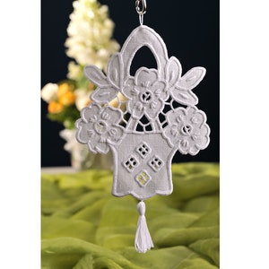 Può includere: Cestino di fiori ricamato bianco con un fiocco che pende dalla parte inferiore. Il cestino è realizzato in tessuto bianco e presenta un delicato motivo a pizzo.