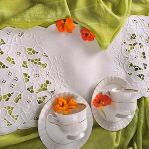 May include: White lace table runner featuring intricate floral embroidery. Two white teacups and saucers with gold trim are positioned on the runner, surrounded by orange blossoms.
