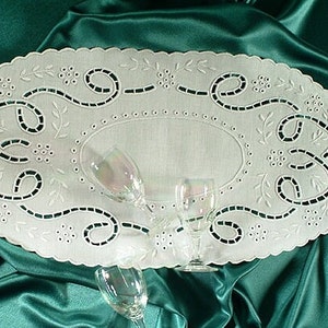 May include: White oval embroidered table runner with a scalloped edge. The runner has an openwork design with floral and scroll patterns. Three clear wine glasses are arranged on the runner, set against a teal satin background.