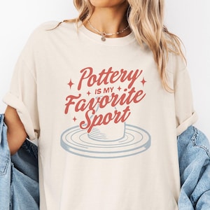 May include: A beige t-shirt with the text "Pottery is my favorite sport" in red lettering. A stylized image of a pottery wheel is below the text.