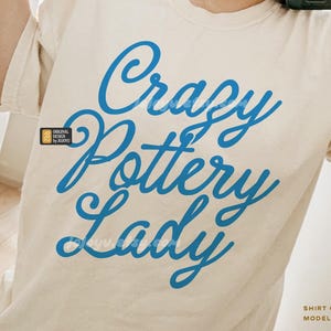 May include: A white t-shirt with the text "Crazy Pottery Lady" in blue cursive lettering. The shirt is being modeled by a person wearing a size XL.