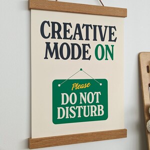 May include: A beige canvas print with a wooden frame and a hanging cord. The print features the text "CREATIVE MODE ON" in black and green lettering. Below the text is a green sign with white lettering that reads "Please DO NOT DISTURB".
