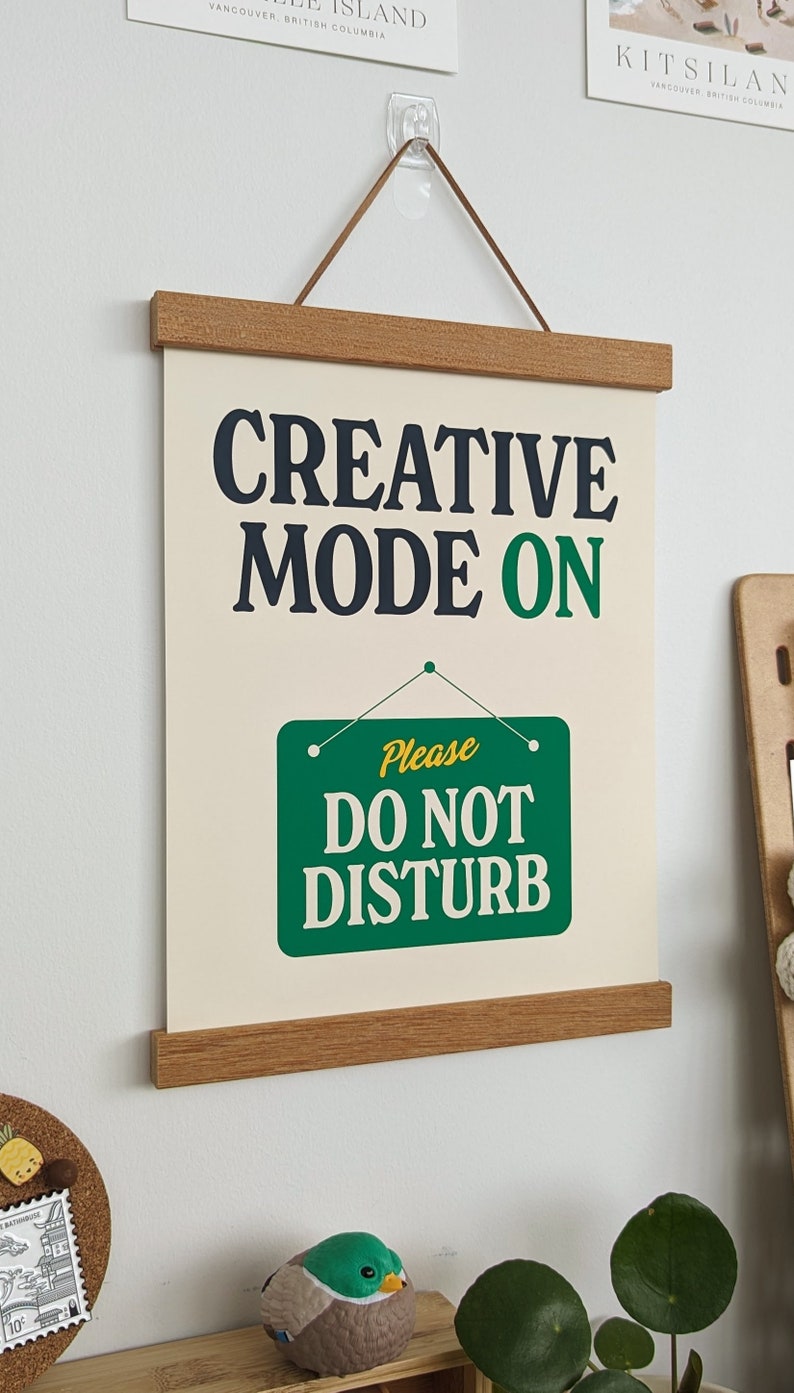 May include: A beige canvas print with a wooden frame and a hanging cord. The print features the text "CREATIVE MODE ON" in black and green lettering. Below the text is a green sign with white lettering that reads "Please DO NOT DISTURB".