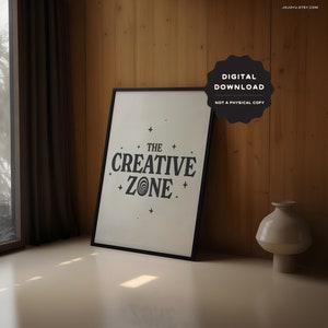 May include: A black framed print with a white background and black text that reads "The Creative Zone" with a stylized swirl design in the word "Zone". The print is on a wooden surface with a vase next to it.