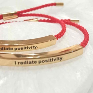 Peut inclure: Deux bracelets en cordon rouge avec des barres dorées qui disent "Je rayonne de positivité."