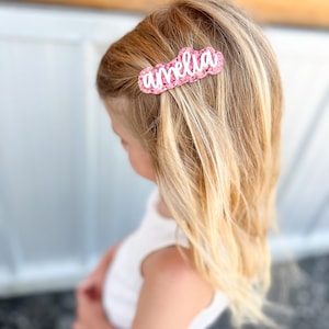 Pu&ograve; includere: Un fermaglio per capelli glitter rosa con il nome "amelia" scritto in lettere bianche.