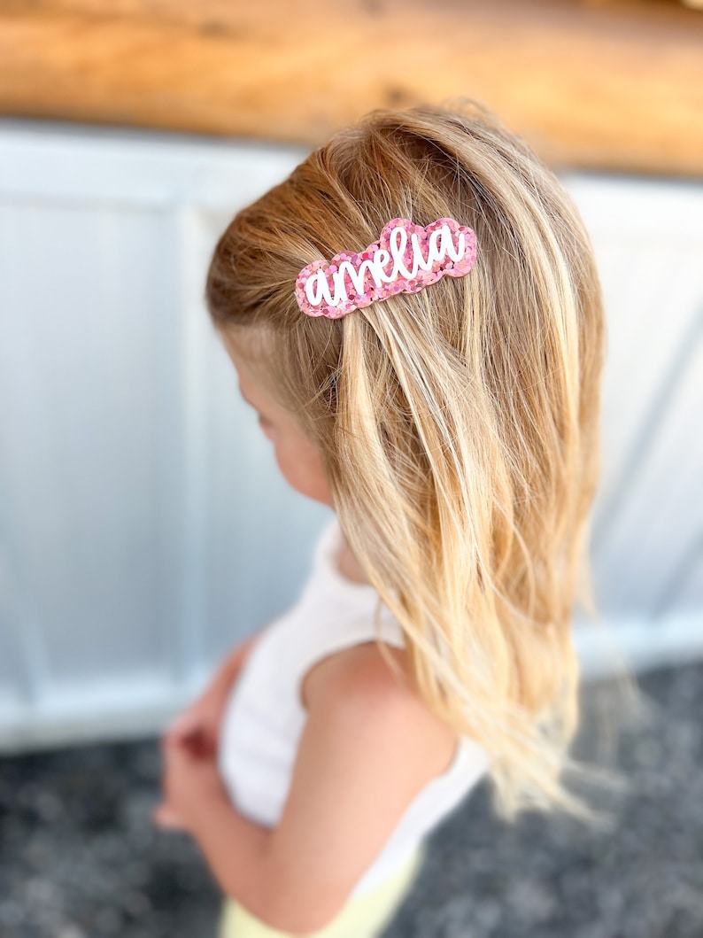 Pu&ograve; includere: Un fermaglio per capelli glitter rosa con il nome "amelia" scritto in lettere bianche.