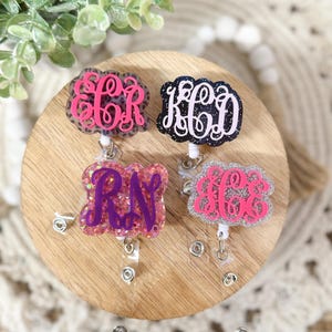 May include: Four monogram badge reels in pink, black, and purple. Each reel features a different initial combination in a decorative font, set against a glittery or solid background. The reels are attached to a clear plastic retractable cord.