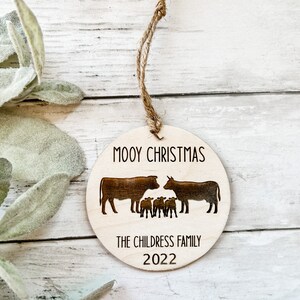 Peut inclure: Une décoration de Noël en bois avec un motif de vache et de veau. L'ornement est gravé avec le texte "Mooy Christmas" et "The Childress Family 2022".