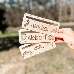 Könnte beinhalten: Drei Holz-Lesezeichen mit verschiedenen Designs und Namen. Das obere Lesezeichen hat ein Blumendesign und den Namen "amelia". Das mittlere Lesezeichen hat ein Fuchs-Design, der ein Buch liest, und den Namen "Abbott". Das untere Lesezeichen hat ein Regenbogen-Design und den Namen "Lyla".