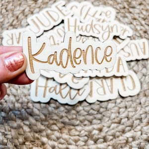 May include: A collection of wooden name tags with various names including "Kadence", "Jude", "Claire", "Hudsyn", and "Hallie". The tags are made of light wood with a natural finish and have a slightly rounded shape.