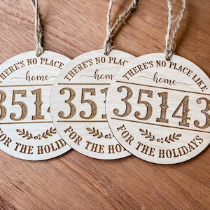 Op de afbeelding: Drie ronde houten ornamenten met touwhangers. Elk ornament heeft de tekst "THERE'S NO PLACE LIKE HOME" en een postcode. De zin "FOR THE HOLIDAYS" is ook opgenomen. De ornamenten zijn lichtbruin.
