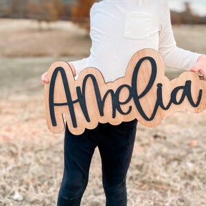 Könnte beinhalten: Ein Holzschild mit dem Namen "Amelia" in schwarzer, kursiv geschriebener Schrift. Das Schild wird von einer Person gehalten, die ein weißes Langarmshirt und eine schwarze Hose trägt.