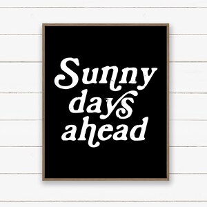 May include: A framed art print with the words "Sunny days ahead" in white, stylized font on a black background. The frame is a natural wood color. The artwork is displayed against a white, horizontal-planked wall.