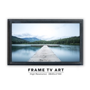 May include: A framed photograph of a wooden dock extending into a still lake with a mountain range in the background. The water is a light blue color and the sky is a light gray color. The frame is a dark gray color.