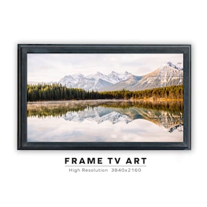 May include: A framed photograph of a mountain lake with snow-capped peaks reflected in the water. The lake is surrounded by a forest of evergreen trees. The sky is a light blue with some clouds.