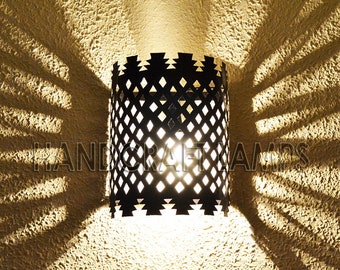 May include: A black metal wall sconce with a geometric diamond pattern and a decorative top edge. The sconce casts a patterned light and shadow on the textured wall. The text "HANDCRAFT LAMPS" is visible.