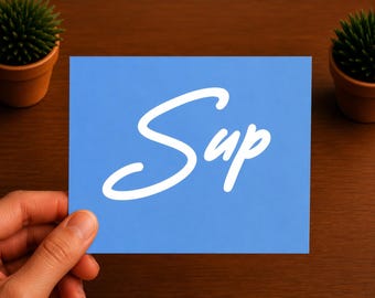 Puede incluir: Una tarjeta rectangular de color azul claro con la palabra Sup escrita en una fuente cursiva blanca. La tarjeta se sostiene sobre una superficie marrón oscuro con dos pequeñas plantas verdes en macetas en las esquinas superiores.