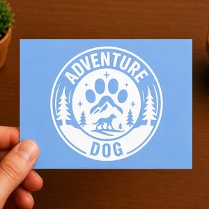 Puede incluir: Una tarjeta rectangular de color azul claro con un gráfico circular blanco. El diseño incluye una huella sobre una cordillera y la silueta de un perro, rodeados de pinos, estrellas y el texto ADVENTURE DOG en un borde circular.