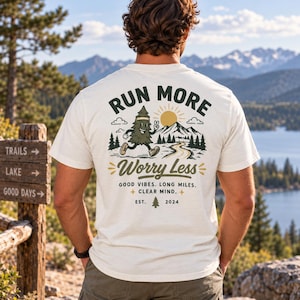 May include: An off-white t-shirt featuring a graphic on the back with a cartoon pine tree running, mountains, and the text: RUN MORE Worry Less, GOOD VIBES. LONG MILES. CLEAR MIND. EST. 2024. The design includes a sun, clouds, and trees.