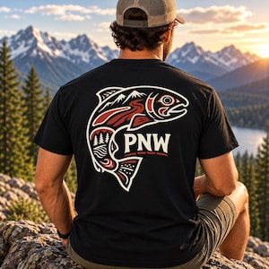 Op de afbeelding: Een zwart t-shirt met korte mouwen en een grote afbeelding op de achterkant van een gestileerde vis gevuld met berg-, bos- en rivierafbeeldingen. De tekst PNW staat in het wit in het midden van het visontwerp met rode en witte accenten.