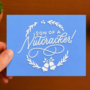 May include: A rectangular light blue greeting card featuring white text that reads Son of a Nutcracker! in a mix of block and script fonts. The text is framed by a white circular wreath design with holly leaves, berries, and two small bells.