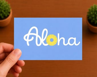 Puede incluir: Una tarjeta rectangular de color azul claro con el texto cursivo blanco Aloha, donde la letra o se sustituye por un gráfico de rodaja de piña amarilla. El diseño está centrado en la tarjeta, que tiene un acabado mate y liso.