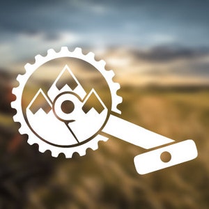 Könnte beinhalten: Weißes Mountainbike-Kurbelgarnitur-Logo mit einem Bergpanorama in einem Zahnrad. Das Logo vor einem verschwommenen Hintergrund aus Feld und Himmel, das Outdoor-Abenteuer und Radfahren suggeriert.