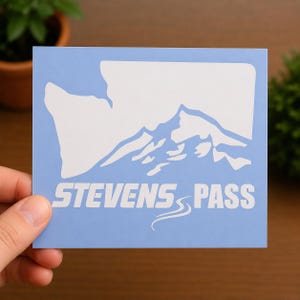 May include: A light blue rectangular sticker featuring a white graphic of a mountain range silhouette and the text STEVENS PASS in a bold, white, sans-serif font with a small white curved line graphic underneath the word PASS.
