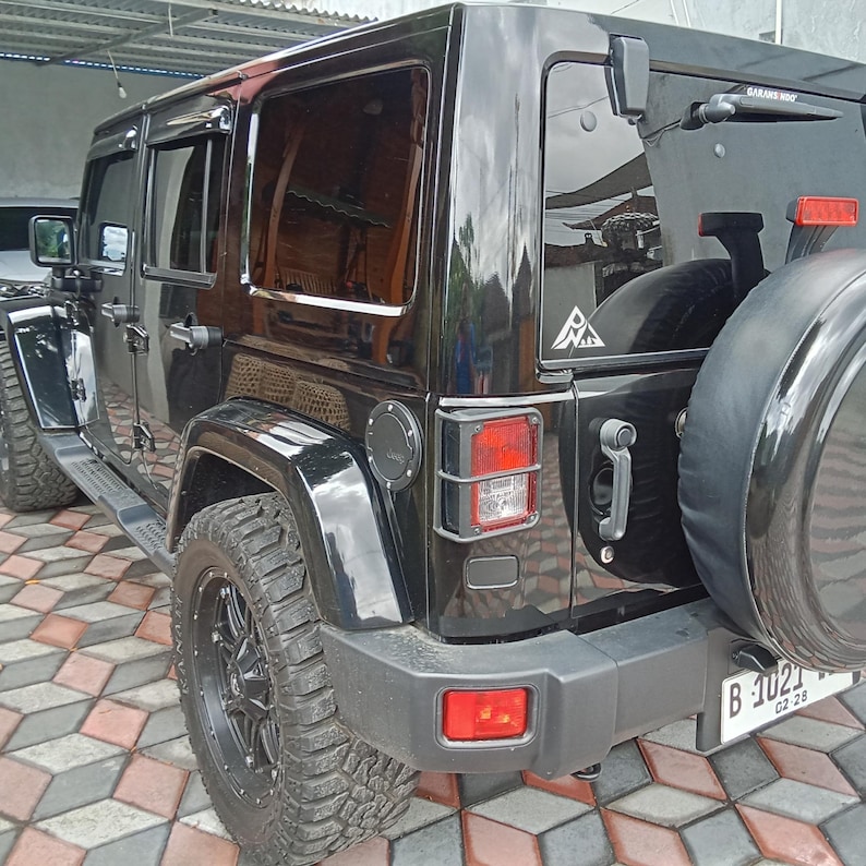 May include: A black four-door SUV with tinted windows, rugged off-road tires, and a rear-mounted spare tire. The vehicle features a square taillight with a protective black cage, a rear wiper, and a license plate reading B 1021.