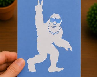 May include: A rectangular blue card featuring a white die-cut silhouette of a yeti wearing sunglasses. The yeti is depicted in a standing position with one arm raised, displaying a peace sign gesture with its fingers, while facing forward.