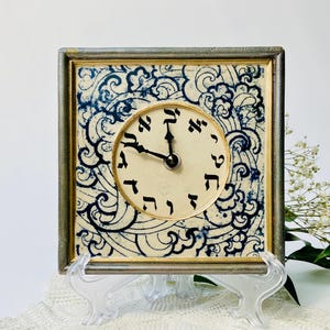 May include: Square clock with a cream-colored face and black hands, set within a silver frame. The clock face features Hebrew numerals. The background has a blue and white swirling pattern, adding a decorative touch to the timepiece.