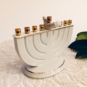 May include: White ceramic menorah with a gold trim and a dove figurine on top. The menorah has nine candle holders and is designed in a traditional style.