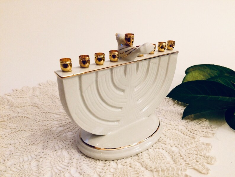 May include: White ceramic menorah with a gold trim and a dove figurine on top. The menorah has nine candle holders and is designed in a traditional style.